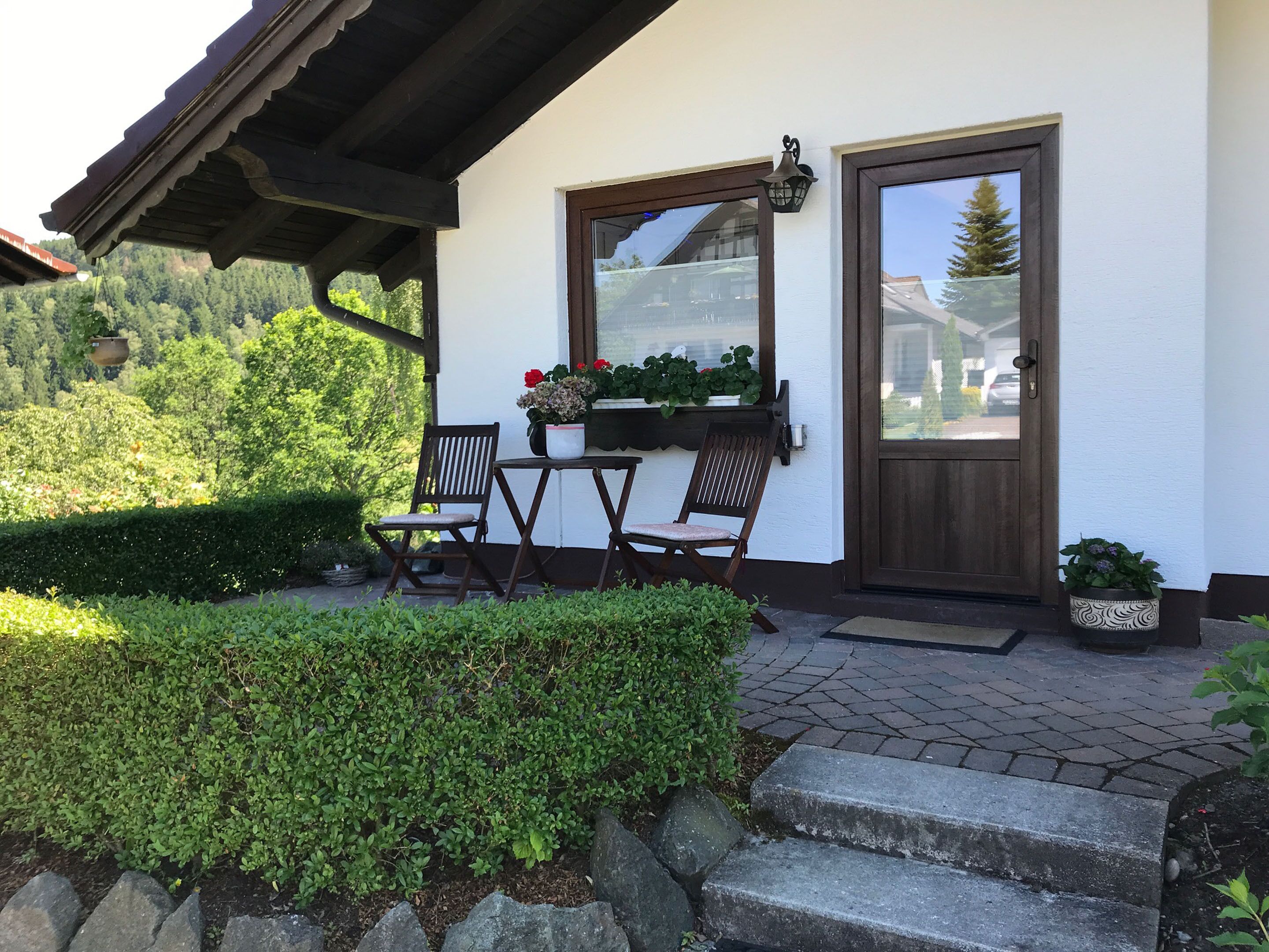 Ein überdachter Balkon mit zwei Holzstühlen und einem Tisch, eine Tür mit Fenster sowie ein Blumentopf auf den Stufen.