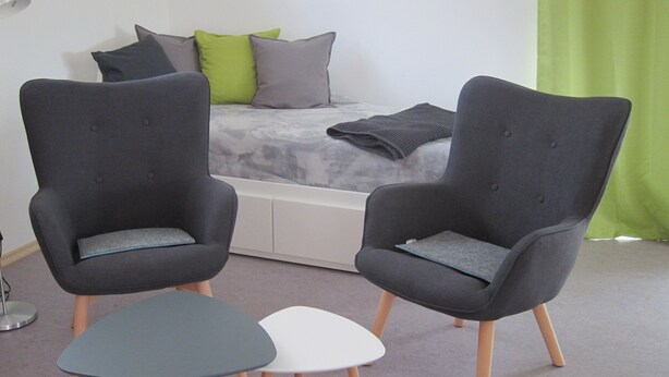 A bedroom with two grey armchairs, a white side table, and a bed with grey and green pillows.