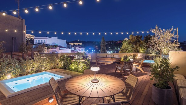 Une terrasse sur le toit avec un jacuzzi, des meubles en bois, des guirlandes lumineuses et une vue sur les bâtiments en arrière-plan.