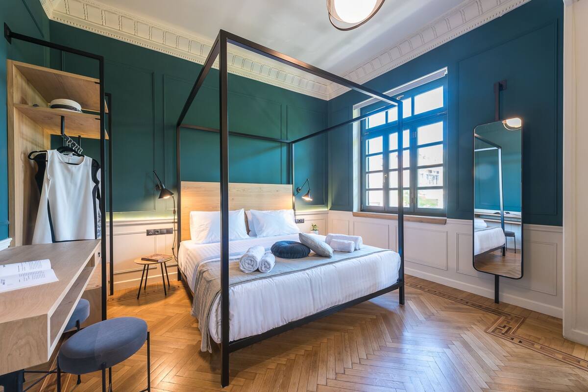 A modern bedroom with a four-poster bed, a desk with a chair, and a large window providing natural light.
