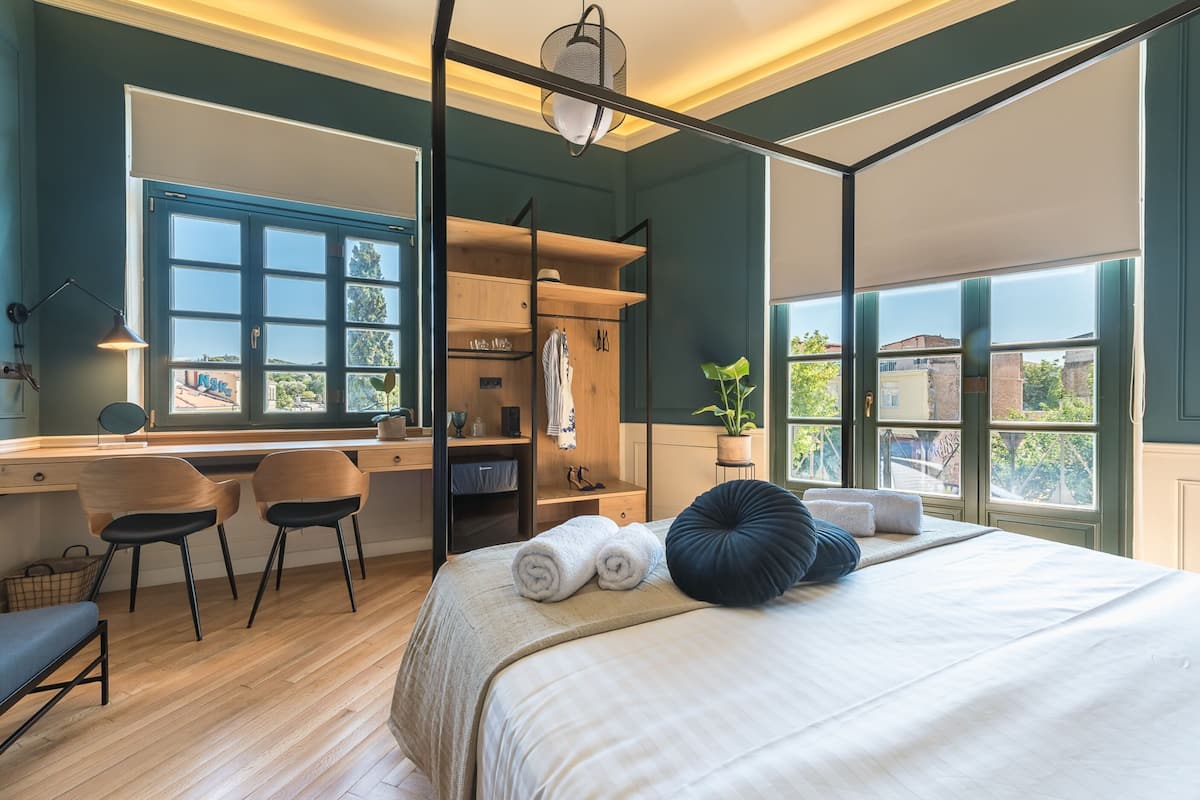 A bedroom with a large bed, a desk, and a chair. There is a window with a view of buildings outside.