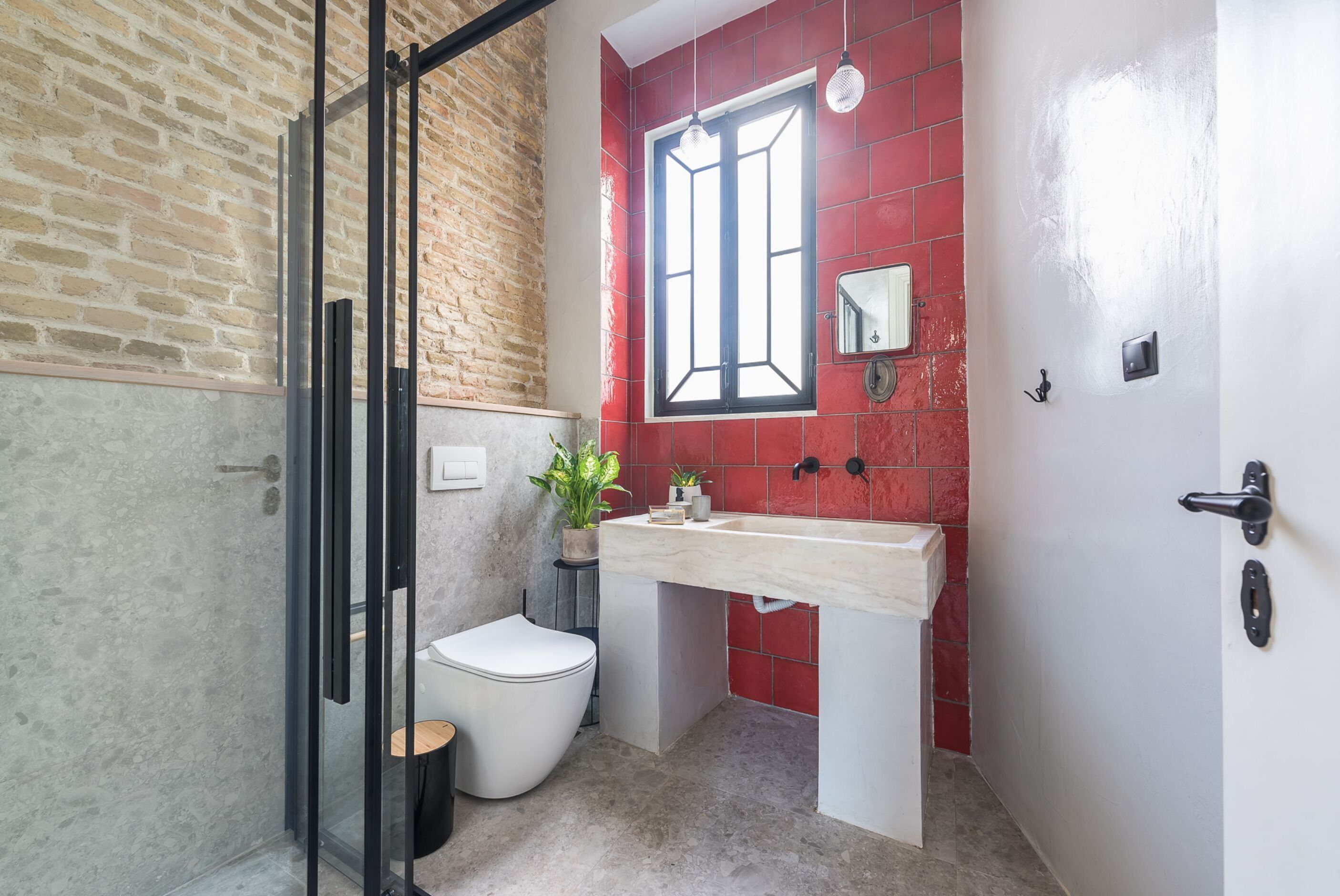 Une salle de bain avec une douche à parois de verre, des toilettes blanches, un lavabo avec un miroir et un mur recouvert de carreaux rouges.