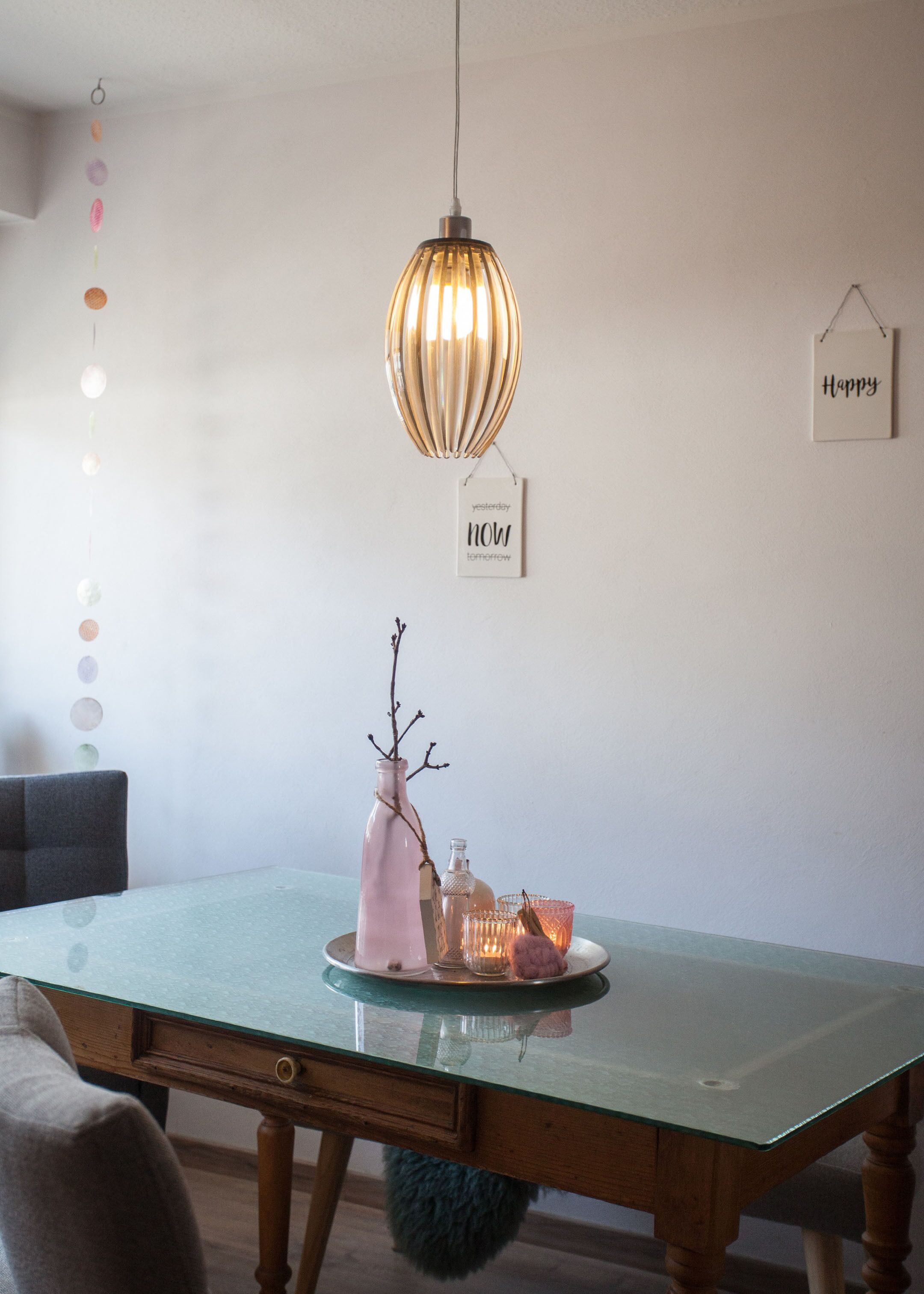 Uma área de refeições com mesa de vidro, luminária pendente, um vaso com galhos e um castiçal.