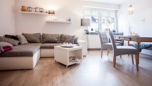 A modern living room with a sectional sofa, a glass-top coffee table, and a dining area with a chair and a lamp. - Ferienwohnung Mia (Winterberg)