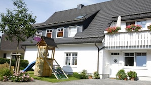 A residential building with a white exterior, a wooden play structure, and a garden area with flowers and a tree. - Ferienwohnung Mia (Winterberg)