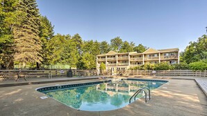 Une piscine entourée d’une terrasse, un immeuble à plusieurs étages doté de balcons, et une forêt dense en arrière-plan.