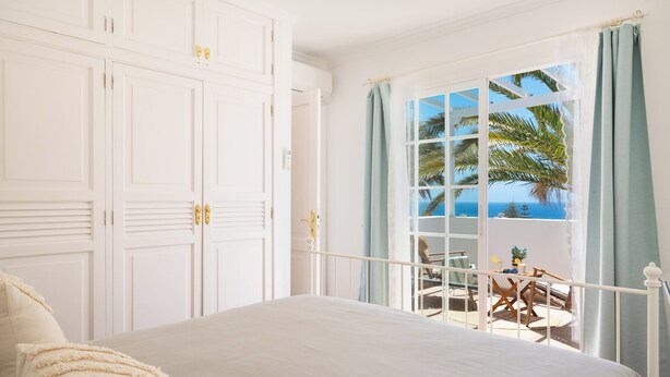 Un dormitorio con una cama grande, un armario y un balcón con vistas al mar y palmeras.