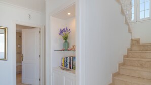 Un pasillo con una escalera de mármol, una puerta blanca y un estante con libros y un jarrón de flores.