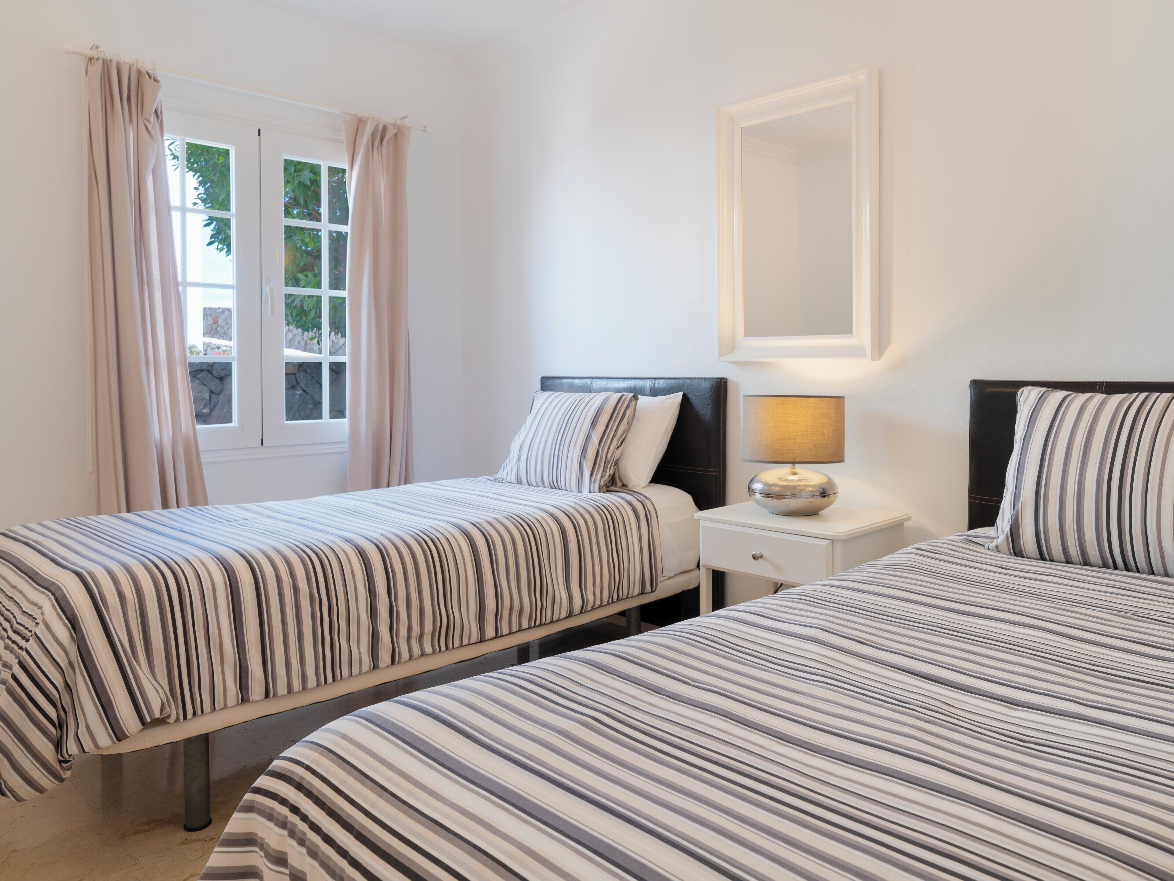 Ein Hotelzimmer mit zwei Betten, einem Nachttisch mit Lampe, einem Fenster mit Vorhängen und einem gerahmten Spiegel an der Wand.
