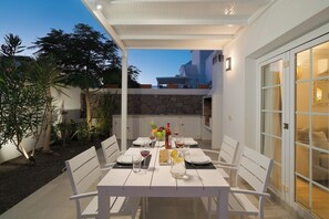 Un área de comedor exterior bien iluminada, con una mesa dispuesta para cuatro personas, rodeada de sillas blancas y un muro de piedra al fondo.