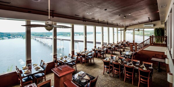 A restaurant with a view of a river and bridge, featuring multiple dining tables and chairs.