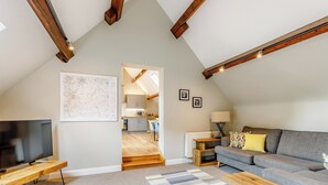 A spacious loft apartment with exposed wooden beams, a large TV, a grey sofa, and a kitchen in the background. - Tadpole Cottage at Frog Hall (Tilston, near Malpas)