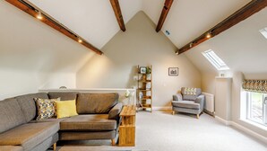 A loft-style living room with exposed wooden beams, a grey sofa, a patterned rug, and a wooden bookshelf. - Tadpole Cottage at Frog Hall (Tilston, near Malpas)
