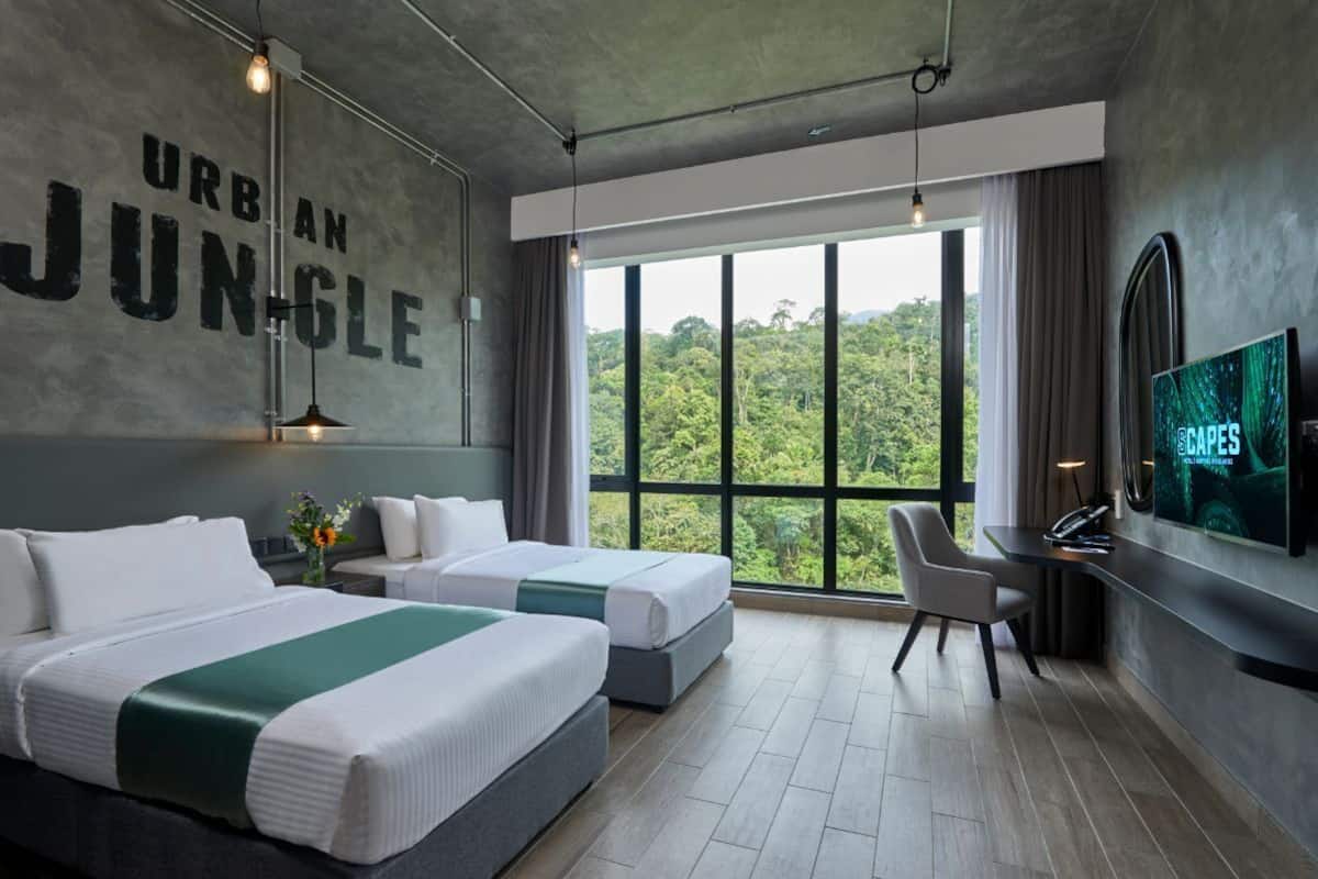 Habitación de hotel con dos camas, un escritorio, televisión y una ventana grande con vistas a la selva.