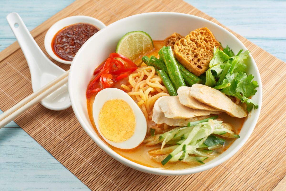 A bowl of noodle soup with various toppings, including vegetables and a boiled egg, served on a white plate with a spoon and a small bowl of sauce.