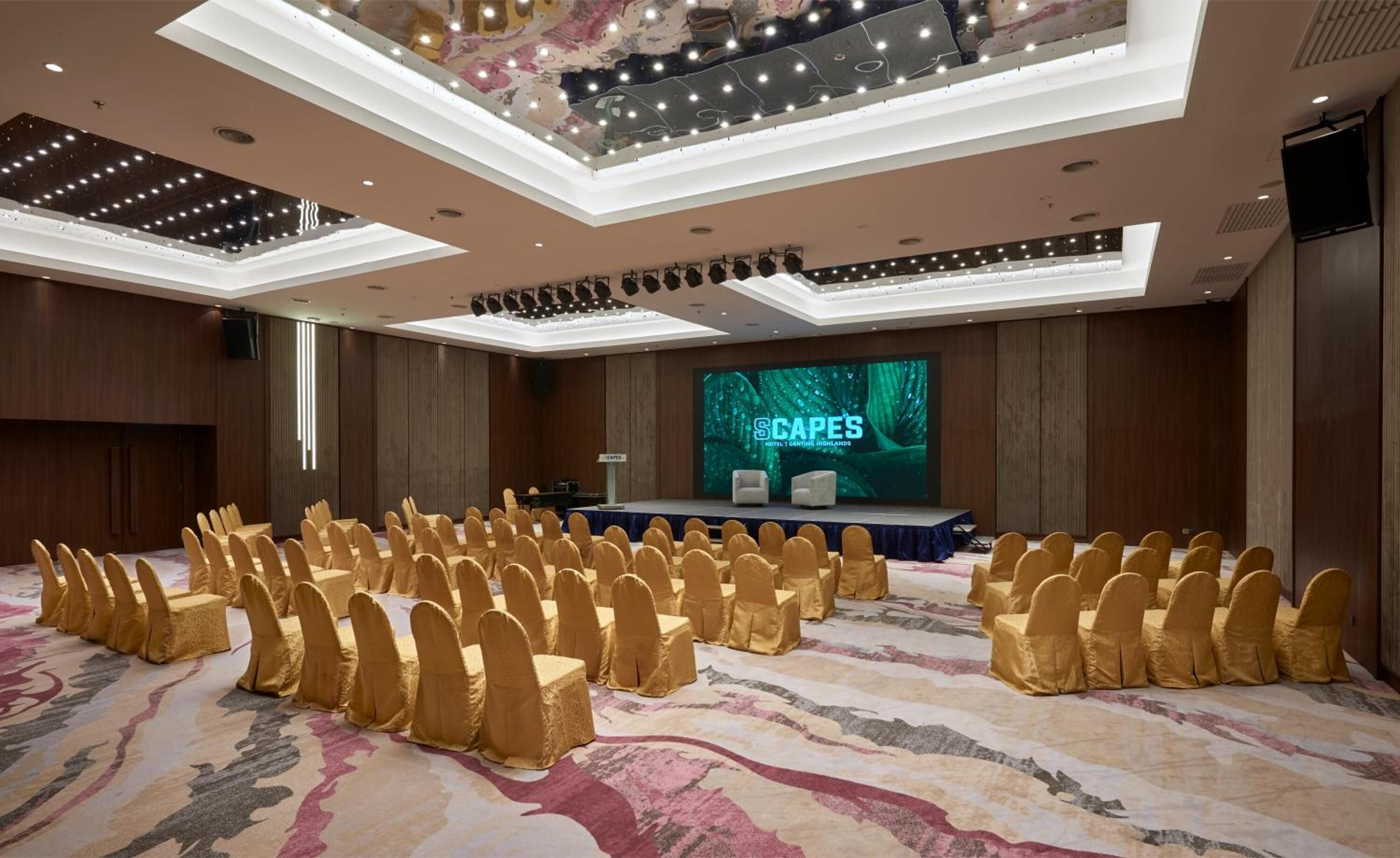 A conference room with rows of chairs, a stage with a screen displaying 'SCAPES HOTEL GENTING HIGHLANDS', and a modern ceiling design.