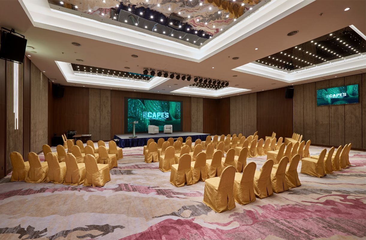 A conference room with rows of chairs, a stage with a screen displaying 'CAPES', and a modern ceiling design.