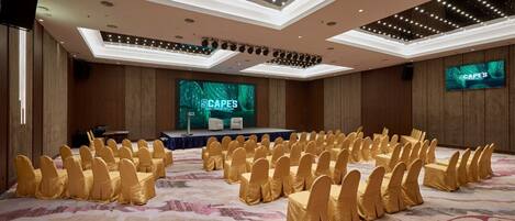 A conference room with rows of chairs, a stage with a screen displaying 'CAPES', and a modern ceiling design.