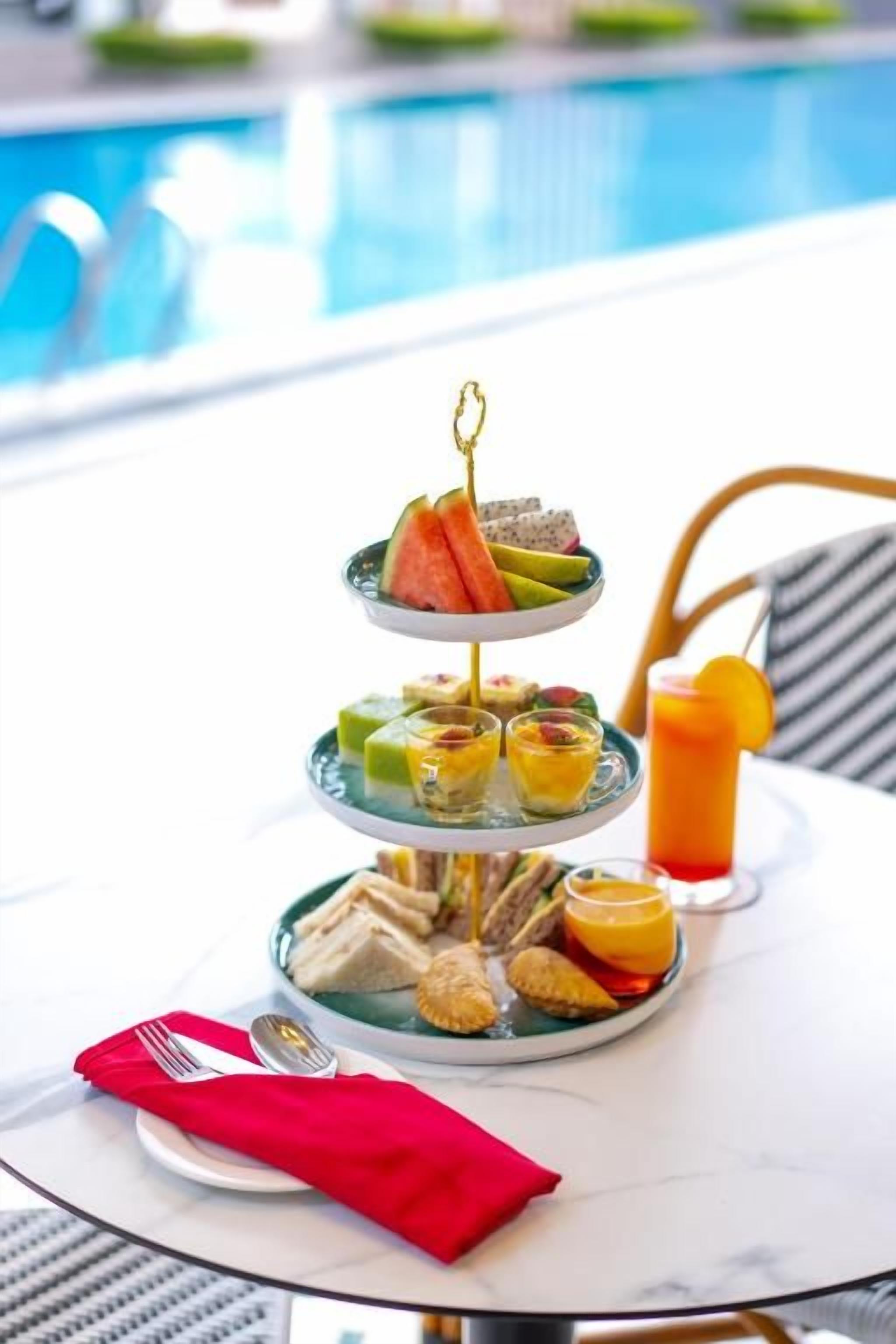 A three-tiered dessert tray with various sweets and a glass of orange juice on a table by a pool.
