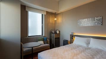 A hotel room with a bed, a seating area with a table, a window, and a wall-mounted light fixture.