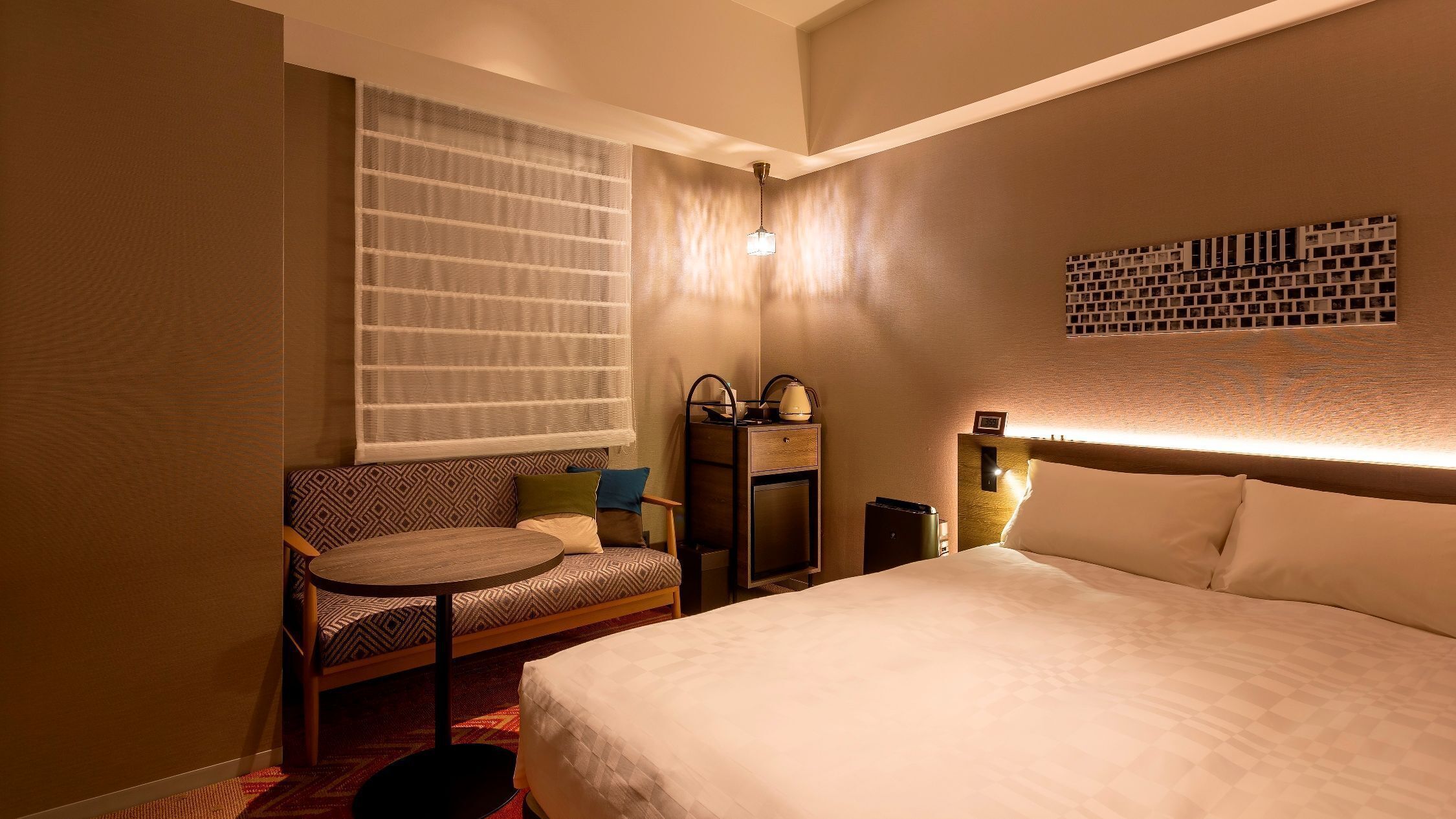 Een hotelkamer met een bed, een stoel, een klein tafeltje, een nachtkastje, een lamp en een raam met dunne gordijnen.