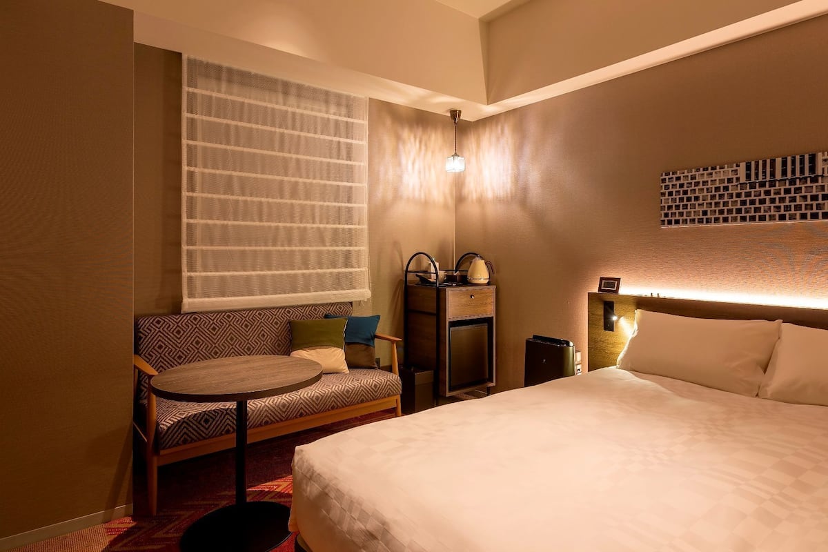 Une chambre d’hôtel avec un lit, une chaise, une petite table, une table de chevet, une lampe et une fenêtre avec des rideaux légers.