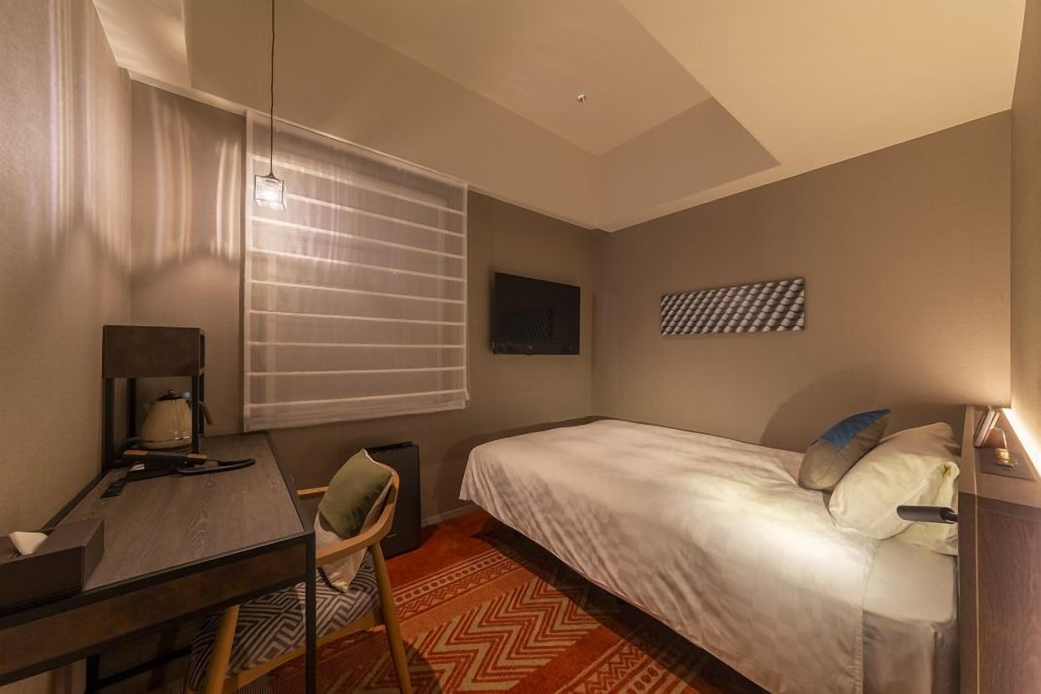 Een hotelkamer met een bed, een bureau, een stoel, een raam met jaloezieën, een aan de muur bevestigde televisie en een kamerbreed tapijt met een patroon.