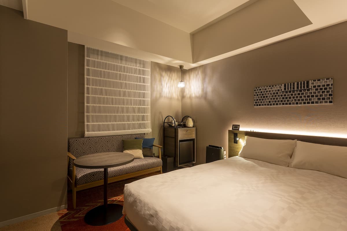 Une chambre d’hôtel comprenant un lit, un canapé, une petite table, une table de chevet, une lampe fixée au mur et une fenêtre avec des stores.