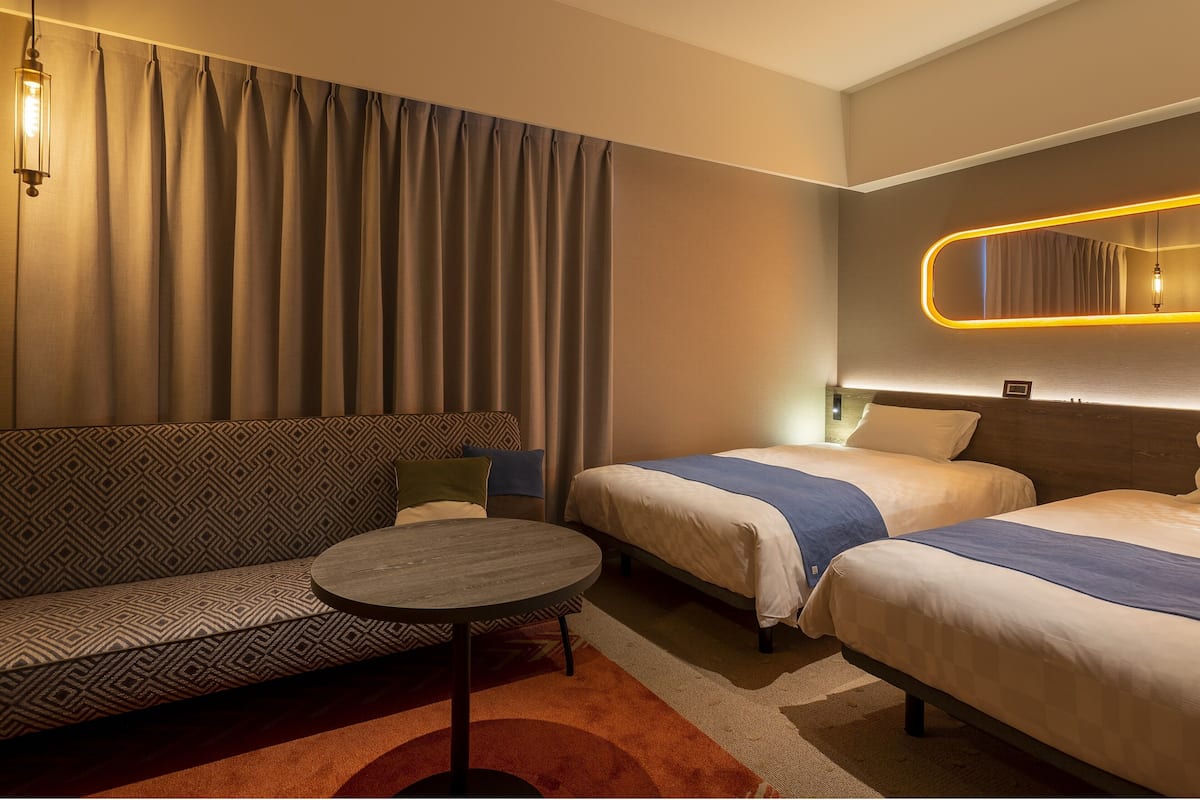 Une chambre d’hôtel avec un canapé, deux lits simples, une petite table ronde et un miroir avec un éclairage intégré.