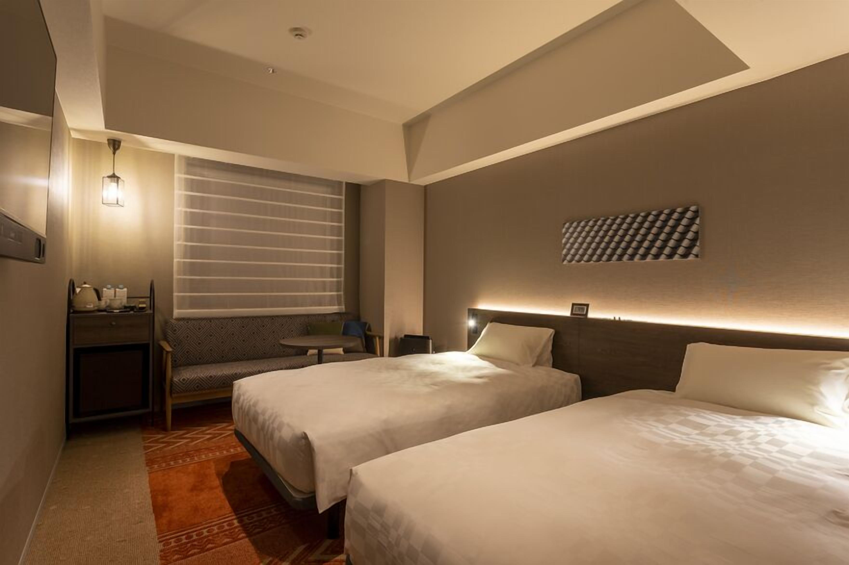 Een hotelkamer met twee bedden, een bureau, een stoel, een kleine tafel, een wandlamp en een raam met zonwering.