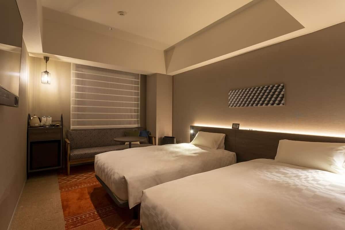 Une chambre d’hôtel avec deux lits, un bureau, une chaise, une petite table, une lampe fixée au mur et une fenêtre avec des stores.