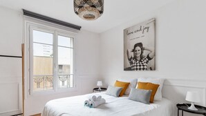 Et hotelværelse med en stor seng, hvidt sengetøj, et vindue med udsigt, et billede på væggen og et lille natbord med en lampe.