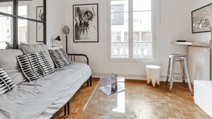 Et moderne, minimalistisk soveværelse med en stor seng, en lille skammel, en radiator og et vindue med udsigt til bygninger.