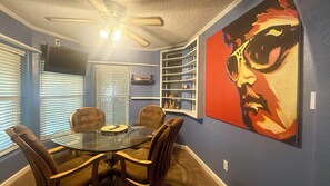 A dining area with a glass table and chairs, a ceiling fan, and a large painting on the wall. - 3-Bed 2 Bath Cozy Foothills Vacation Home (Yuma)