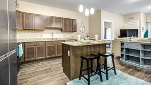 A modern kitchen with wooden cabinets, a central island with stools, and a dining area with a grey console table. - Clubhouse Fully Equipped Apartment (Mankato)