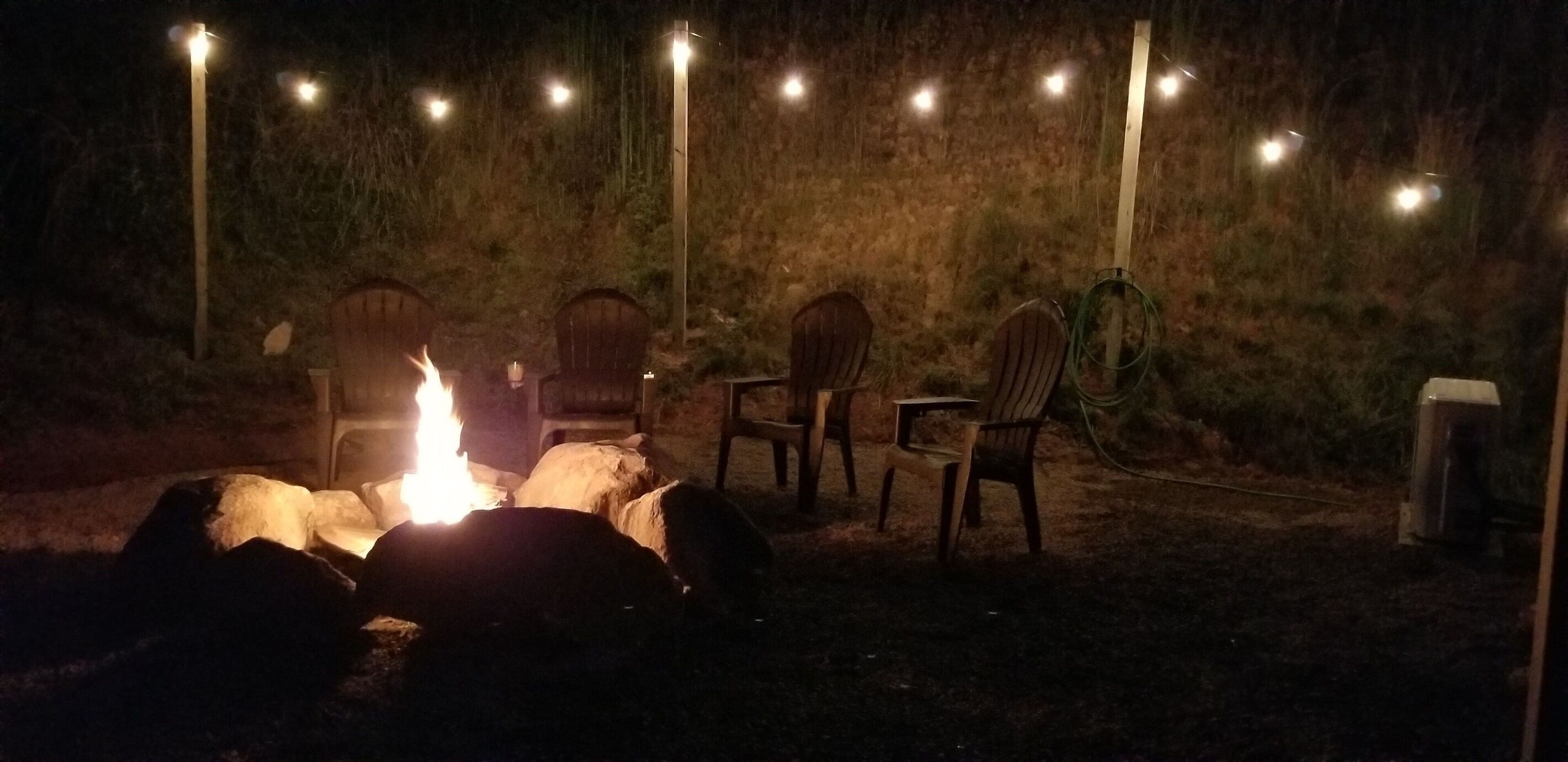 Nuotiopaikka, jossa on tuoleja ja koristeellisia valonauhoja, metsäisellä alueella.