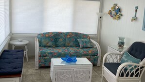 Una habitación iluminada por el sol, con techo blanco, sofá floral, sillón de mimbre, una mesita con un jarrón azul y un banco.