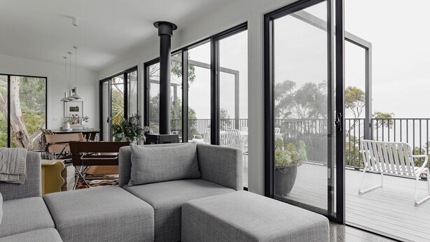 A modern living room with a sectional sofa, a dining area with a table and chairs, and large sliding glass doors leading to an outdoor deck. - Months and Seasons - Coastal House - Stunning Views (Separation Creek)