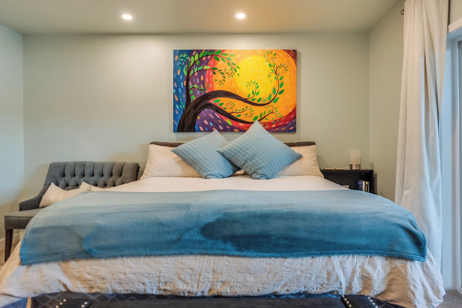 A bedroom with a bed, pillows, a nightstand, a painting, and a window with curtains.