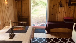 Interior de una cabaña de madera con una cama, una mesa con una vela, un perchero y una puerta que da al exterior.
