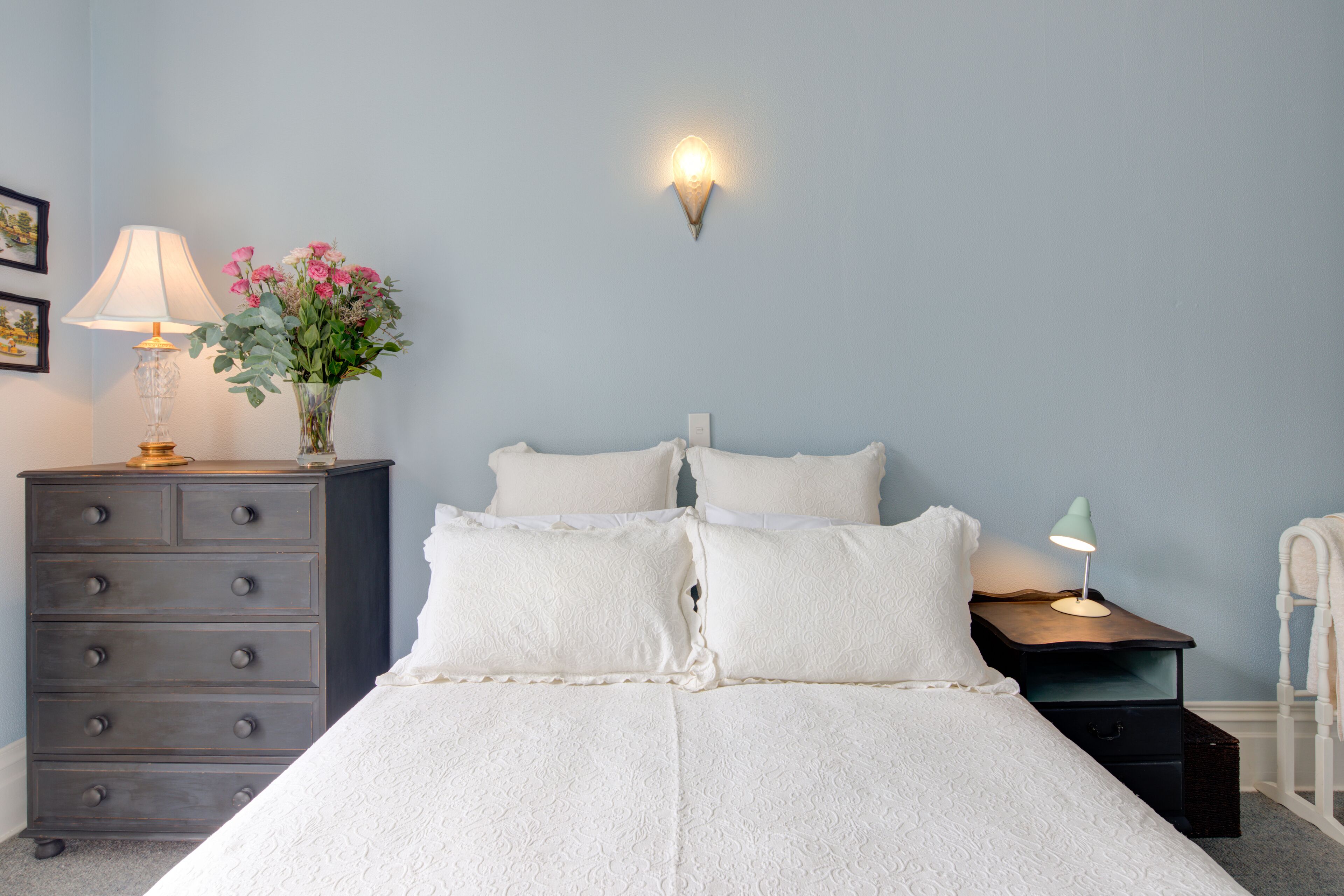 Un dormitorio con una cama, un aparador, una mesita de noche, una lámpara de pared y un florero con flores.