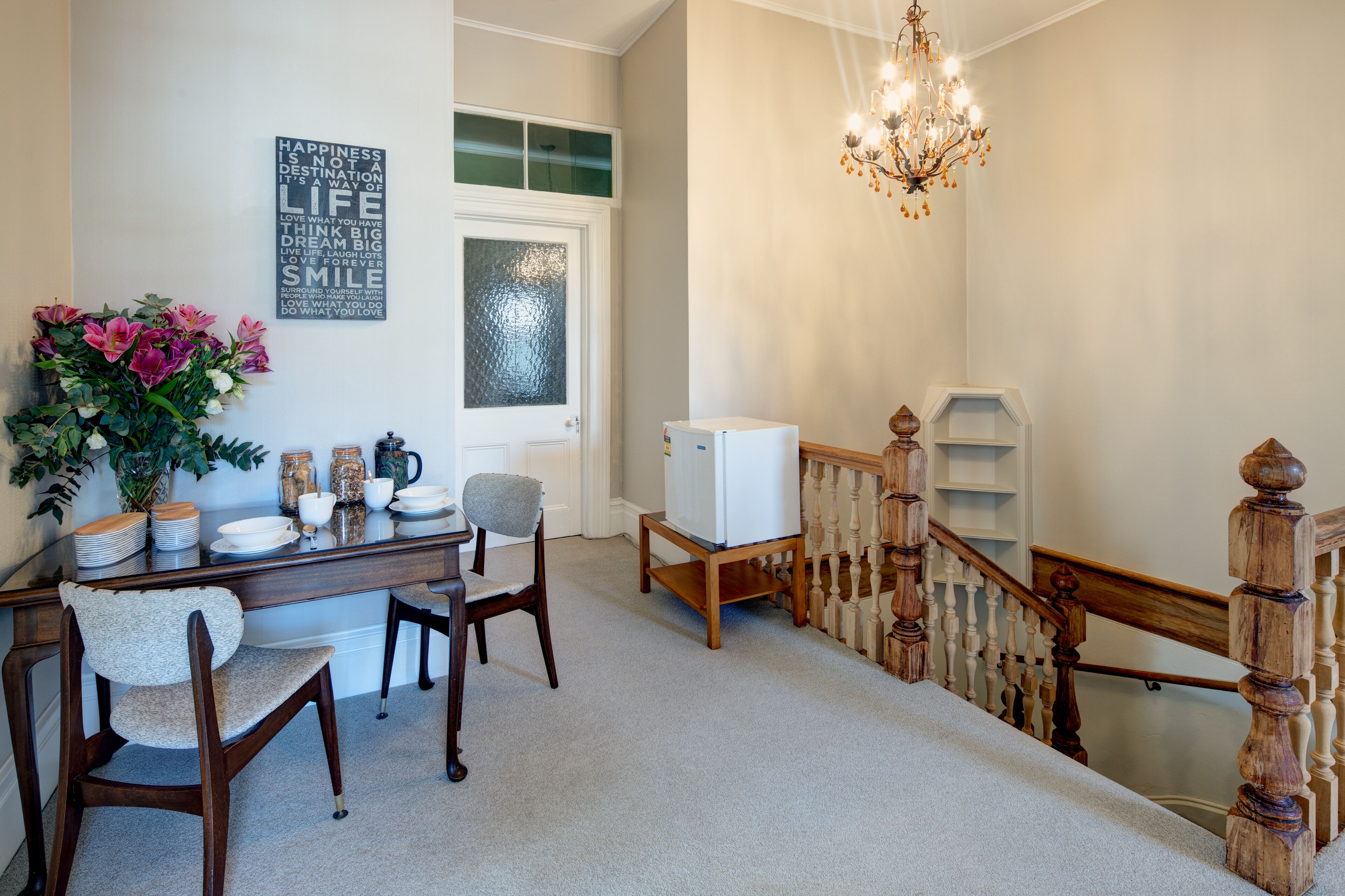 A dining area with a wooden table, chairs, and a refrigerator. A staircase with wooden railings leads to another level.