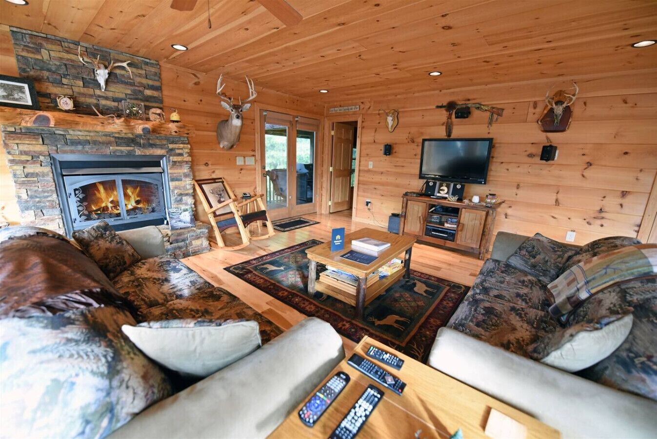 A cozy cabin interior with a stone fireplace, wooden walls, mounted animal heads, a television, and a comfortable seating area.