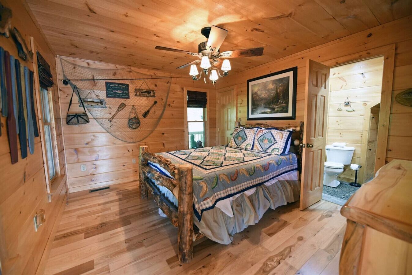 A wooden cabin bedroom with a bed, a ceiling fan, a painting, and a fishing net on the wall.