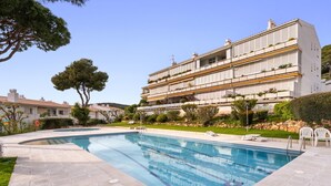 Una piscina con solárium empedrado, un edificio de varios pisos con balcones y un jardín cuidado.