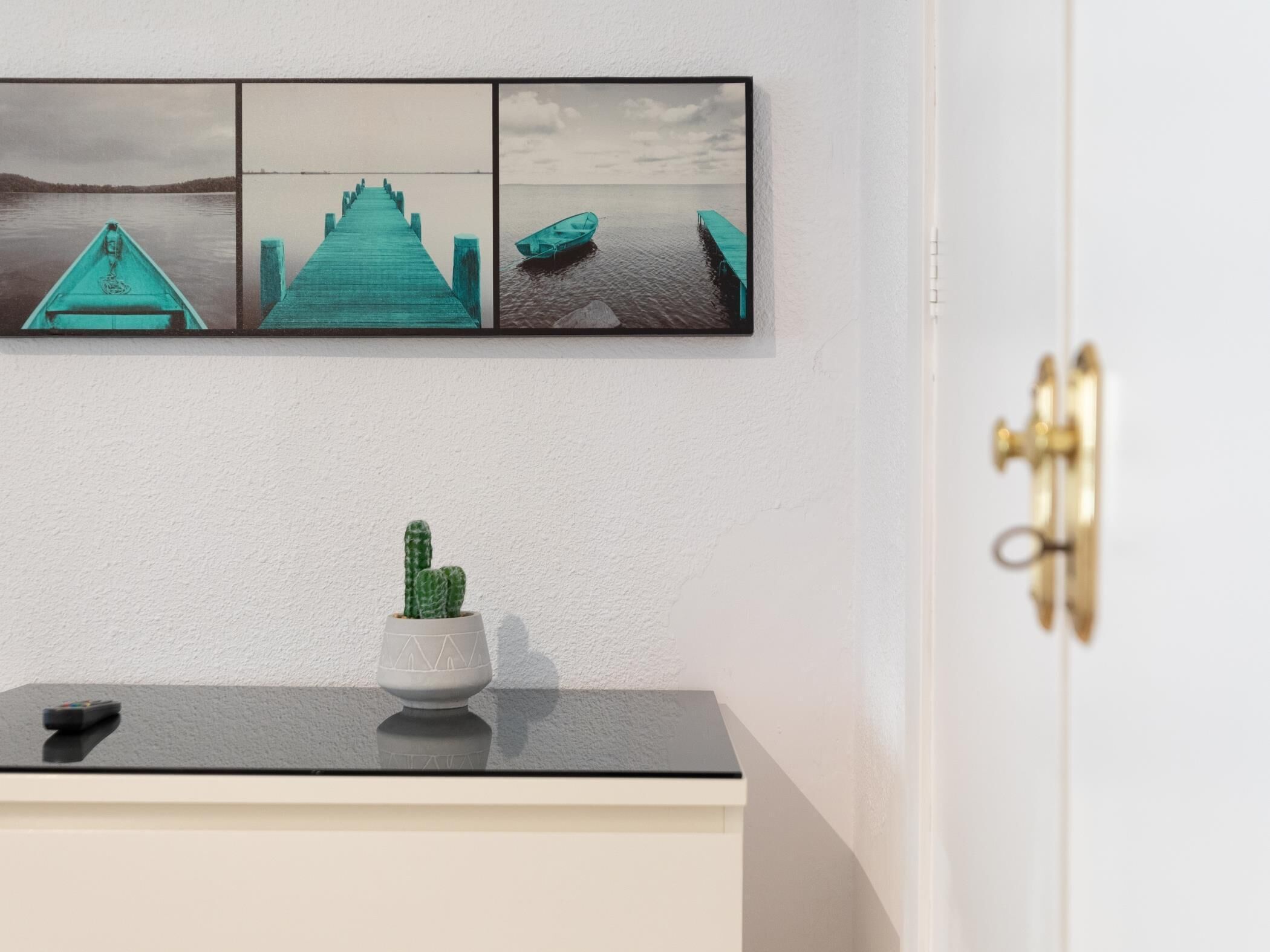 Una obra de arte colgada en la pared que muestra un muelle color turquesa que se extiende sobre aguas tranquilas, una pequeña planta de cactus en maceta y una puerta con tirador de latón.