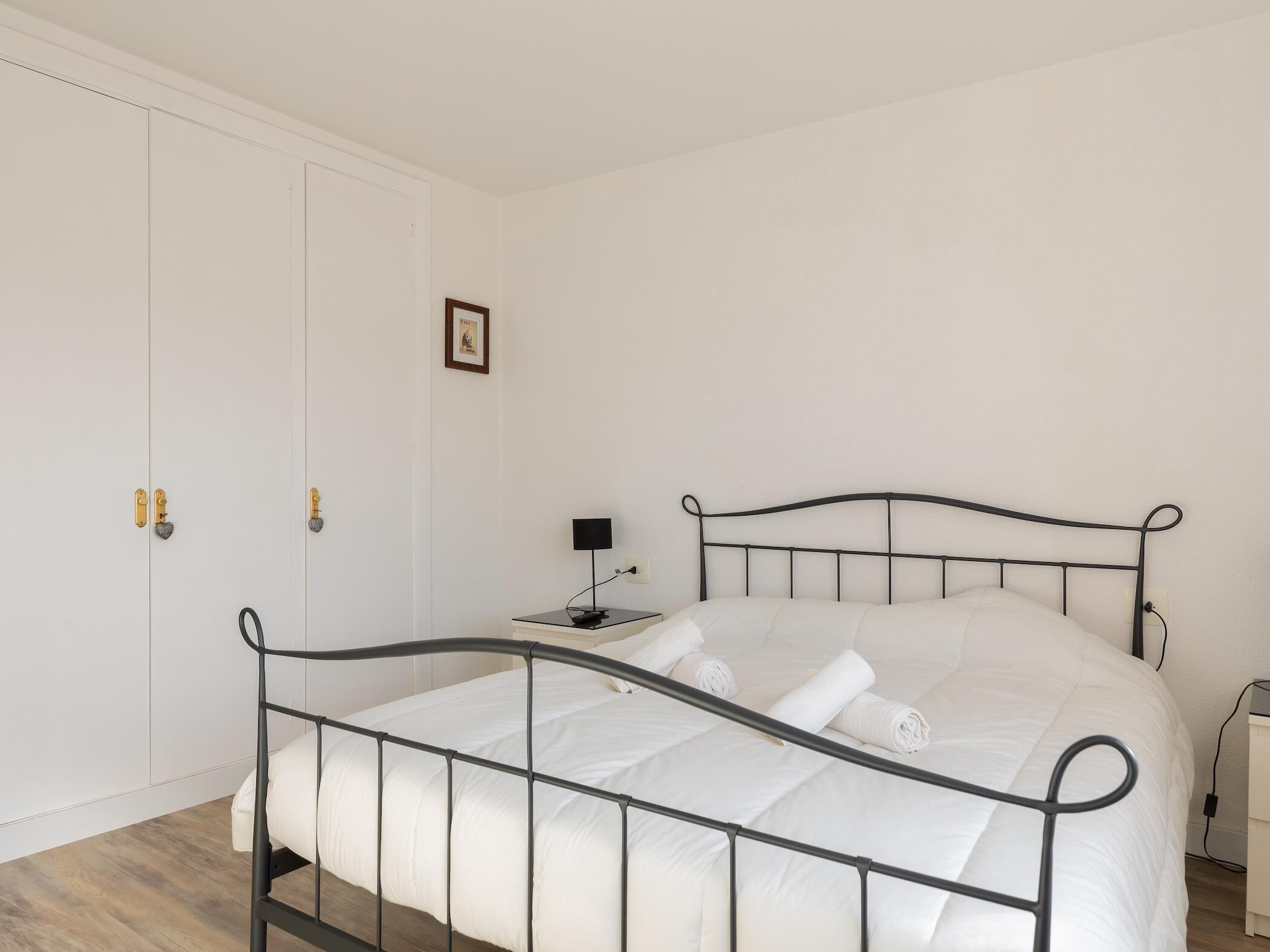 Un dormitorio con estructura de cama metálica, ropa de cama blanca, una mesita de noche con lámpara y una puerta con cerradura.