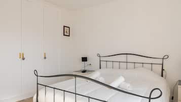 Un dormitorio con estructura de cama metálica, ropa de cama blanca, una mesita de noche con lámpara y una puerta con cerradura.