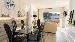 Un espace repas moderne avec une table en verre et des chaises noires, un mur en bois orné d’une horloge représentant une carte du monde, et un balcon aménagé avec du mobilier d’extérieur.