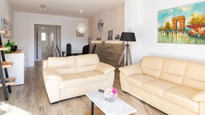 Un salon moderne avec deux canapés beige, un coin repas agrémenté d’un mur en bois et un tableau représentant l’Arc de Triomphe.
